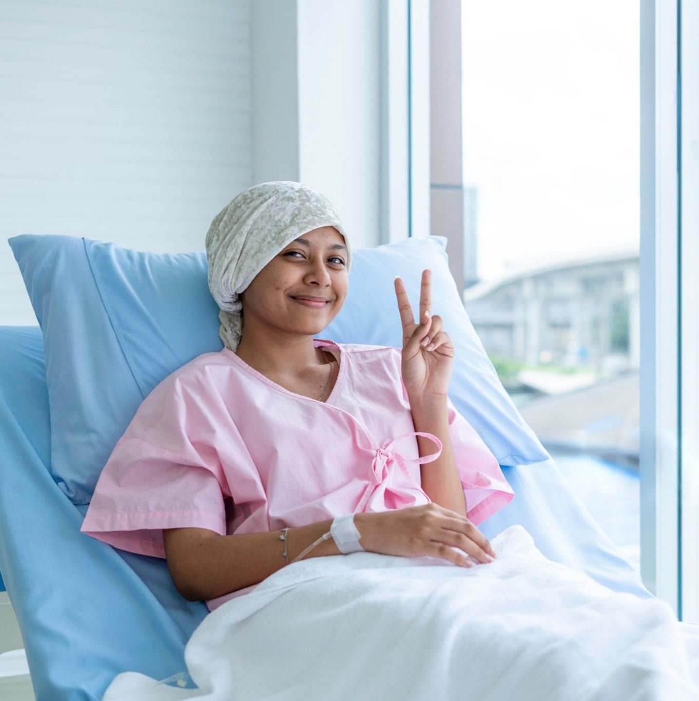 A young cancer patient smiling and showing a victory sign from her hospital bed